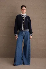 Cardigan de lana color marrón con detalles de bordes en color rojo. Presenta cierre frontal con botones, mangas largas con puños ajustados y un pequeño bordado decorativo en el pecho.