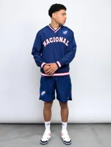 Buzo deportivo azul marino de nylon con cuello en V, puños y cintura acanalados con detalles tricolor (blanco, rojo y azul). Estampado frontal con la palabra "NACIONAL" en blanco y escudo del club C.N.de F. en el pecho. Corte clásico.