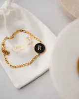 Pulsera dorada con cadena de eslabones y dije redondo negro con la letra R en dorado.