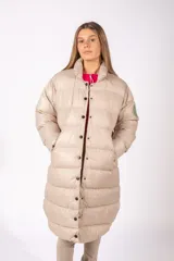 Campera puffer larga de color beige, con cuello alto, cierre frontal con botones a presión y diseño acolchado horizontal. Cuenta con un parche circular verde en la manga izquierda.