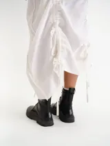 Vestido midi blanco sin mangas, con cuello redondo y diseño holgado. Presenta un cordón ajustable fruncido en el centro de la parte frontal y parches circulares sutiles en la tela.