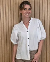 Camisa blanca de algodón fino, de manga corta abullonada con puños elásticos, cuello mao con escote en V y cierre frontal de botones oscuros.