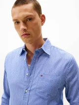 Camisa de hombre de manga larga, confeccionada en sarga de algodón mercerizado, con diseño de rayas verticales azules y blancas. Presenta cuello con botones, bolsillo de parche en el pecho con logo bordado de Tommy Hilfiger y puños redondeados con botón.