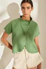 Blusa de lino verde de manga corta y corte recto, con cuello alto con volado y cierre frontal cruzado con botoncitos dorados.