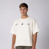 Remera boxy de color crudo con hombro caído y corte cuadrado a la cadera. En el frente presenta un diseño minimalista con texto y un pequeño logo central.