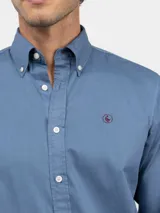Camisa de hombre color azul, con cuello buttondown, cierre frontal con botones y logo bordado en el pecho. Presenta un acabado vintage logrado mediante el proceso de teñido en prenda.