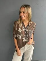 Blusa de corte holgado color chocolate con estampado floral en tonos claros. Presenta cuello camisero con tapeta frontal decorada con lentejuelas y un bolsillo en el pecho también con aplique de lentejuelas. Posee mangas tres cuartos con detalle de trabilla para arremangar.