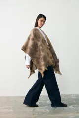 Ruana de tejido de punto con diseño teñido artesanalmente en tonos beige y marrón, con terminación de flecos en el ruedo y etiqueta de cuero sintético en el lateral.