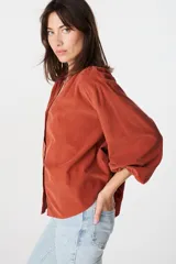 Camisa de corte holgado y diseño minimalista, color crudo, con cuello mao, cierre frontal de botones y mangas largas abullonadas con puños elásticos.