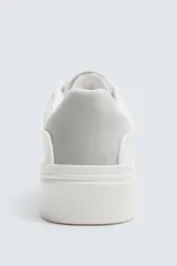 Zapatilla deportiva de cuero sintético color blanco, con suela de plataforma gruesa y detalle de gamuza gris en el talón.