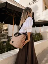 Cartera tipo tote de diseño capitoneado en color marrón claro, con doble asa de mano negra y bolsillos laterales. Presenta un diseño acolchado con líneas verticales en el frente y rombos en los laterales, e incluye una etiqueta decorativa con logo.