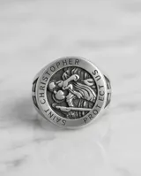 Anillo de acero con diseño circular grabado que representa a San Cristóbal, santo patrono de los viajeros, con detalles en relieve en los laterales.