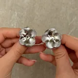 Par de aros tipo caravanas con diseño de flor de cuatro pétalos con textura estriada en acabado plateado.