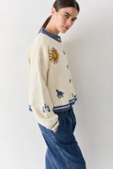 Cardigan de lana color crema con cuello redondo, cierre frontal de botones y mangas largas con puños ajustados. Presenta bordados decorativos en el frente que incluyen un sol, olas y estrellas, con detalles en azul en el cuello y el ruedo.