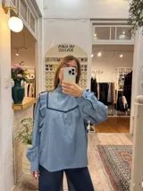 Blusa de jean fino con cuello alto, mangas largas con volumen y volados decorativos en los hombros. Presenta detalles de ribetes en terciopelo azul oscuro en el frente y los volados.