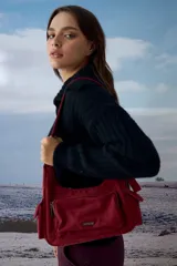 Cartera de hombro color rojo, confeccionada en símil gamuza, con diseño de múltiples bolsillos externos, incluyendo uno frontal con cierre y dos laterales. Cuenta con correa ajustable y logo metálico de la marca en el frente.