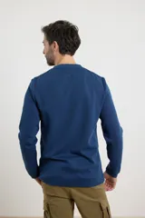 Remera de manga larga en tejido elastizado, color azul, con cuello tipo camisa, cartera con botones y detalles en eco gamuza en la base del cuello y la cartera.