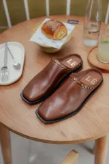 Mules de cuero color suela, con diseño abierto y tira frontal decorada con tachas metálicas circulares. Presentan punta cuadrada y suela plana.