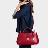 Cartera Guess modelo Carrie de color rojo, confeccionada en símil cuero con herrajes dorados. Presenta doble asa de mano, correa ajustable y desmontable, cierre superior y un detalle de tira frontal con la letra "G" metálica. Incluye un pequeño candado colgante decorativo y el nombre de la marca en la parte inferior.