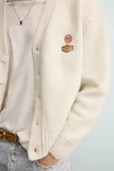 Cárdigan de punto color beige con cuello en pico, manga larga y cierre frontal de botones. Presenta un pequeño bordado a contraste en el pecho y acabados en rib en puños y bajo.