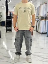 Remera color beige con cuello redondo y mangas cortas. Estampado frontal con el texto "Duxonstudios" en tipografía cursiva, acompañado de un logotipo circular con hojas de laurel y una figura central en tono verde oscuro.