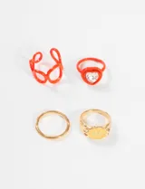 Set de anillos de metal y plástico con talles surtidos. Incluye un anillo dorado con forma de flor y carita sonriente, un anillo dorado liso, un anillo naranja con forma de corazones entrelazados y un anillo naranja con un cristal en forma de corazón.
