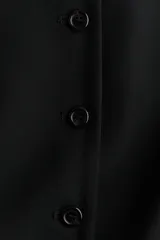 Chaleco largo negro con cuello redondo y cierre de botones frontales. Diseño entallado con bolsillos delanteros ribeteados y abertura en la parte trasera.