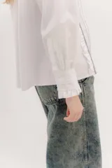 Camisa de algodón blanca con cuello a la base, cierre frontal con botones y mangas largas con puños delicados. Presenta volados verticales y detalles de cintas de terciopelo en contraste a lo largo del frente.