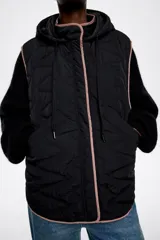 Chaleco acolchado color crema con capucha integrada, cierre frontal de botones a presión y vivos en contraste color negro. Presenta diseño de costuras geométricas y bolsillos laterales.