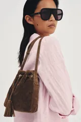 Bolso tipo bandolera de formato mini, confeccionado en piel de ante color marrón. Presenta un diseño de saco con cierre mediante cordón fruncido y detalle de borlas. Incluye una correa larga ajustable y herrajes metálicos dorados.