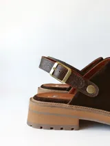 Zueco de cuero gamuzado color chocolate, con diseño de punta cerrada y tira ajustable con hebilla metálica en el talón. Presenta una suela de plataforma dentada en tono marrón claro.