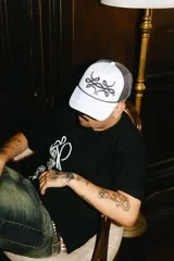 Gorra trucker gris con paneles frontales blancos y paneles traseros de rejilla negra. Presenta un logo bordado en el panel frontal y una cuerda decorativa en la base de la corona.