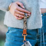 Llavero de muñeca con cinta de tela estampada de camuflaje con patrón de hojas y ramas en tonos marrones y verdes. Tiene un conector de cuero sintético marrón con logo grabado y un gancho metálico color naranja.