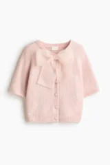 Saco de punto suave color rosado, de manga corta y cuello redondo. Presenta cierre frontal con botones forrados y una moña decorativa de organza transparente en la parte superior. Posee terminaciones acanaladas en mangas y ruedo.