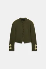 Cazadora militar de color verde oscuro, de cuello subido y manga larga. Cuenta con detalles de alamares y cordones dorados en los hombros y puños, y cierre frontal con botones dorados metálicos.