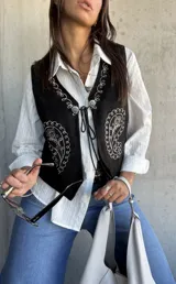Chaleco negro de gamuza con bordados blancos estilo paisley y cierre frontal con cordones y detalles metálicos.