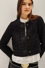 Saco corto de textura tweed en color azul oscuro con destellos metalizados. Presenta cierre frontal mediante tres trabas de cuero sintético con botones tipo toggle, cuello redondo y mangas largas.