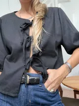 Blusa de lino color negro con cierre frontal mediante tres lazos o moños. Presenta mangas cortas abullonadas con puños elásticos y un corte holgado.