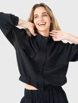 Buzo negro de tela suave y ligera con cuello alto, cierre frontal con botones a presión, bolsillos en la parte frontal y pretina elástica en la cintura.