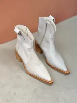 Botas estilo texano de caña baja en cuero color natural, con puntera fina y tacón cuadrado de madera. Presentan costuras decorativas en el empeine y cierre lateral.