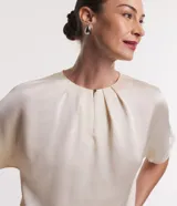 Blusa de manga corta color crema, confeccionada en tejido satinado liso. Presenta un cuello redondo con pliegues tipo canesú en la parte delantera y un pequeño cierre con botón en la nuca.