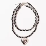 Gargantilla de doble cadena, una de eslabones plateados y otra de cordón trenzado negro, con dije de corazón plateado.