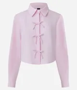 Camisa cropped de manga larga en tejido tricoline, con estampado de rayas verticales finas en color rosa y blanco. Presenta cuello camisero tradicional y cierre frontal decorado con tres lazos de tela. Diseño de corte corto y ajuste recto.