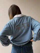 Buzo tipo chomba de corte oversize con diseño a rayas horizontales celestes y blancas. Presenta cuello camisero blanco y un bordado en la espalda con la inscripción República Oriental del Uruguay.