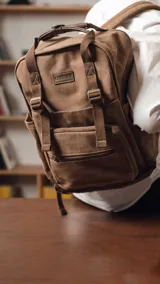 Mochila urbana de tela canvas color marrón, con diseño funcional que incluye compartimento para notebook de 15 pulgadas y espacio interior organizado para set de mate. Cuenta con correas ajustables, manija superior reforzada, bolsillo frontal con cierre y detalles en cuero sintético.