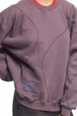 Buzo blanco de manga larga con diseño holgado, cuello redondo y costuras externas visibles en contraste. Cuenta con una etiqueta decorativa en el frente inferior.