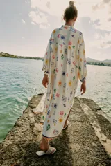 Vestido largo estilo túnica de color crudo con estampado multicolor de motivos marinos y mediterráneos. Presenta cuello con volante, cierre frontal con botones, manga larga y cordón ajustable en el cuello con abalorios.