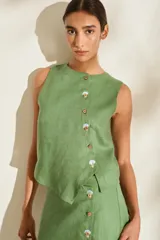 Top de lino blanco sin mangas, con cuello redondo y cierre de botones de madera dispuestos en una cartera asimétrica. Cuenta con delicados detalles bordados en forma de flor alrededor de los ojales.