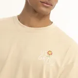 Remera marrón con estampado pequeño en el pecho que dice "We all grow" con una flor.