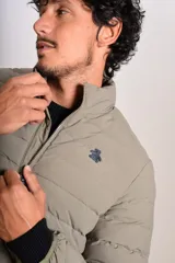 Campera puffer de corte clásico en color beige, con cierre frontal, cuello alto y puños elásticos. Presenta un diseño acolchado horizontal y un pequeño logo bordado en el pecho.